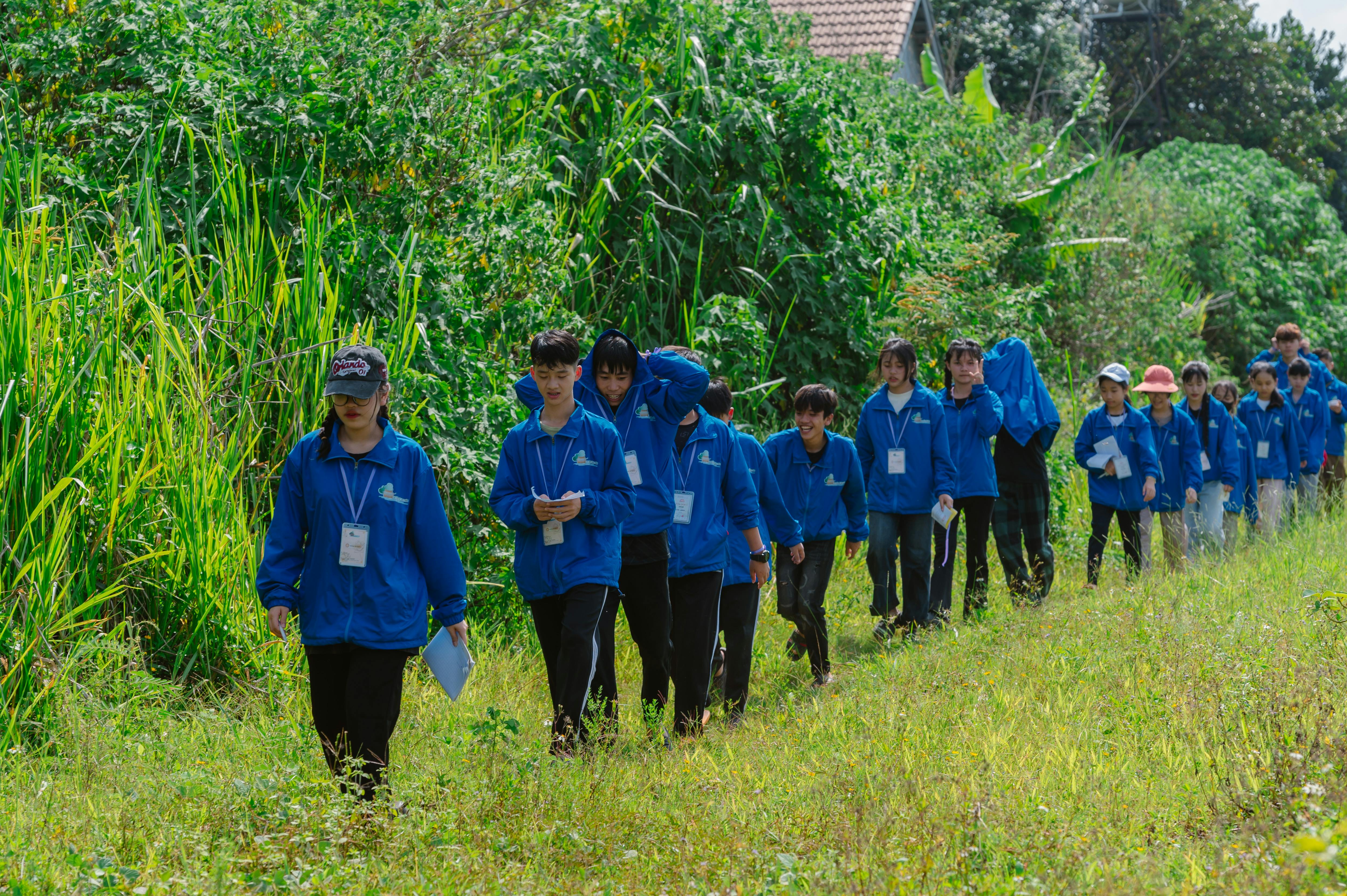 A group of teenagers walks along a grassy path in nature, wearing blue uniforms and carrying papers.